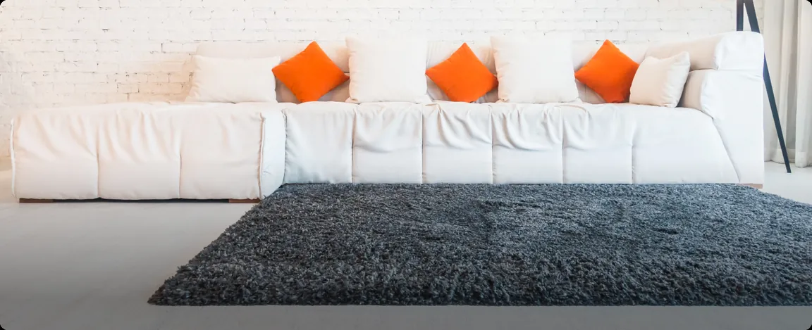 A dark deep-pile carpet in front of a white sofa with orange cushions